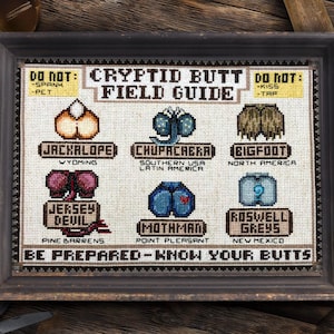 Puede incluir: Obra de arte en punto de cruz enmarcada titulada "Cryptid Butt Field Guide". La obra presenta ilustraciones bordadas de traseros de críptidos, incluyendo Jackalope, Chupacabra y Bigfoot, con etiquetas de texto. La frase "Be Prepared - Know Your Butts" está en la parte inferior.