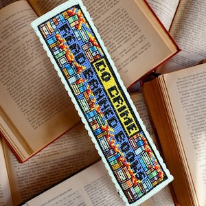 May include: A white bookmark with a colorful cross-stitch design. The bookmark features a stack of books with the text "To Crime I Read Books".