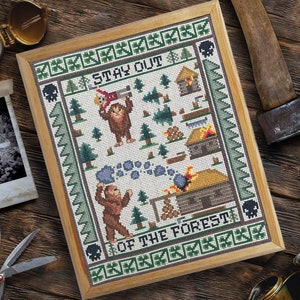 May include: A framed cross-stitch pattern with a green border and a white background. The pattern depicts a cartoon Bigfoot holding a tree and a sign that reads "Stay Out of the Forest".