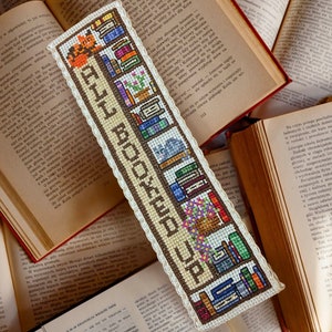 Puede incluir: Un marcapáginas beige con un diseño de punto de cruz marrón y blanco de una estantería con libros, un gato y una planta en maceta. El marcapáginas tiene un borde de ganchillo blanco y el texto "ALL BOOKED UP" en marrón.