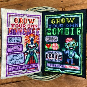 May include: Two cross-stitch samplers with the text "Grow Your Own Banshee" and "Grow Your Own Zombie". The Banshee sampler is white with purple accents and a green and orange figure. The Zombie sampler is black with green accents and a blue and red zombie figure.