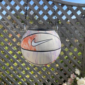 May include: A basketball planter with a white base, a colourful abstract design, and a white Nike logo. It hangs from a black chain. The background includes a wooden trellis and a blue sky. The planter is a creative garden accessory.