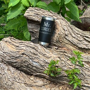 May include: Black stainless steel tumbler with the text "Mr. Cordes - Est 6.5.21" engraved on it. The tumbler is sitting on a tree trunk.