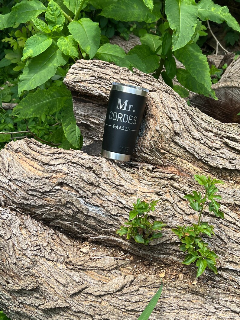 May include: Black stainless steel tumbler with the text "Mr. Cordes - Est 6.5.21" engraved on it. The tumbler is sitting on a tree trunk.