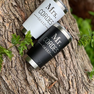 May include: Two stainless steel tumblers with black and white lids. The tumblers are personalized with the text "Mr. Cordes Est 6.5.21" and "Mrs. Cordes Est 6.5.21".