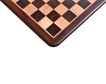 May include: A square wooden chessboard featuring a classic alternating pattern of dark brown and light tan squares. The board is framed by a smooth, dark brown wooden border with rounded corners, showcasing a natural wood grain finish.