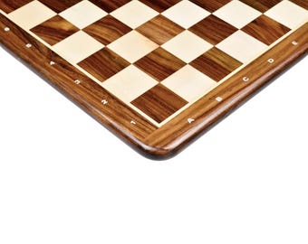 May include: A wooden chessboard featuring a classic alternating pattern of dark brown and light cream-colored squares. The board has a polished wooden border with white alphanumeric coordinates printed along the edges to mark the rows and columns.