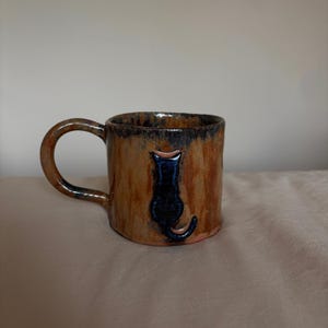 May include: A handmade ceramic mug with a glossy brown and tan mottled glaze. The mug features a dark blue, raised relief silhouette of a cat sitting with its back turned, positioned centrally on the exterior surface, and a curved handle.
