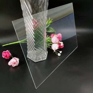 May include: A clear acrylic sheet, also known as plexiglass, is shown on a black surface. The sheet is propped up at an angle, and a clear glass vase with pink flowers is in front of it.