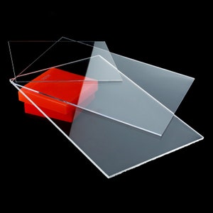 May include: Three clear acrylic sheets stacked on top of each other. The sheets are rectangular and have a slight sheen. The top sheet is slightly smaller than the bottom sheet. The sheets are resting on a red box.