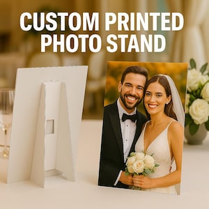 May include: A white custom printed photo stand displaying a photograph of a smiling couple. The photo stand has an easel back. The photograph shows a man in a black tuxedo and a woman in a white wedding dress, holding flowers.