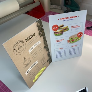 May include: A brown and white menu board with a coffee shop logo and a list of hot and cold coffee drinks. The menu board also includes prices for each drink. The board is on a white surface.