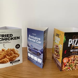 May include: Three tabletop signs, each with a different theme. The first sign advertises fried chicken for £5. The second sign advertises a rental car. The third sign advertises pizza with the text "Delicious Pizza Time" and "Hot & Fresh".