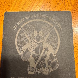 May include: Square, dark gray slate coaster featuring a laser-etched image of Deadpool with crossed swords. The text reads "THE MERC WITH A MOUTH WANTS YOU!" and "JOIN THE FOURTH WALL BREAKERS".