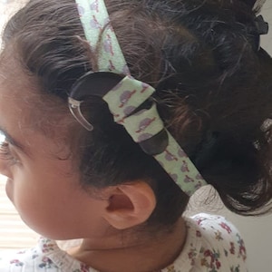 Può includere: Una bambina con i capelli castani indossa una camicia bianca con un motivo floreale. Indossa una fascia verde e bianca con un piccolo fermaglio nero.