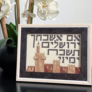 May include: A framed cross-stitch embroidery featuring a cityscape with a tall building and the Hebrew words "ירושלים" (Jerusalem) and "עם ישראל" (People of Israel).