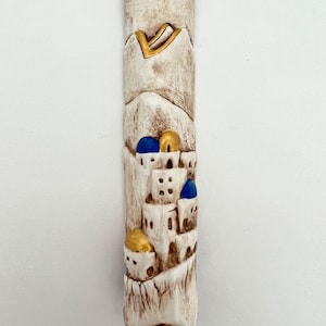 May include: White ceramic mezuzah with an embossed design of a city with gold and blue accents. The design features buildings with blue domes and gold accents. The mezuzah is a traditional Jewish religious item.