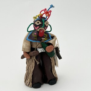 May include: A handmade clay figurine of a person wearing a colorful mask, brown pants, and a green vest. The figurine has a red nose and a colorful string of yarn on its head.