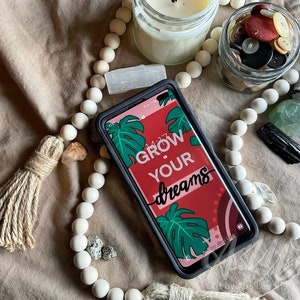 May include: A black phone case with a red, pink, and green tropical leaf design. The case has the text "Grow Your Dreams" in white lettering.