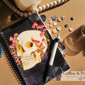 Op de afbeelding: Een zwart spiraalnotitieblok met een schedel-illustratie omringd door rode en gele paddenstoelen. Het notitieblok heeft de tekst "Coffins & Peonies Handmade by Ashlee" in de rechterbenedenhoek.