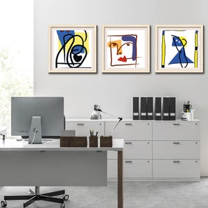 May include: Three framed abstract art prints with geometric shapes and lines in blue, yellow, red, and black. The prints are hung on a white wall in an office setting.