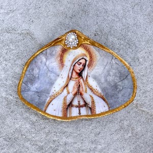 May include: A decorative seashell featuring a decoupage image of the Virgin Mary in a white and gold robe with a halo. The shell has a gold-painted edge and is adorned with a single white pearl surrounded by small beads at the top center.
