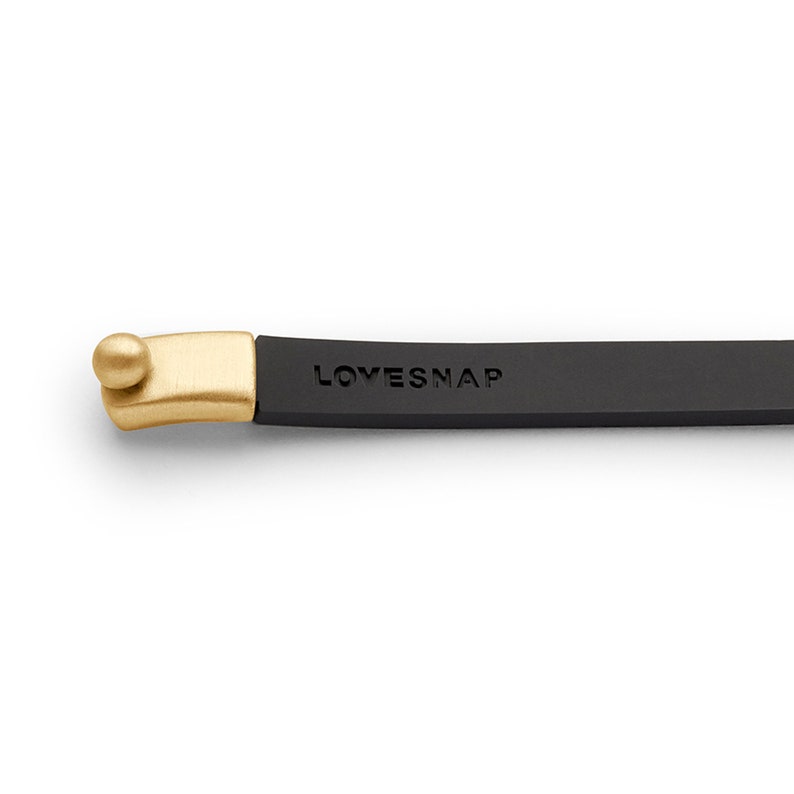 Puede incluir: Un brazalete de cuero negro con un cierre dorado y la palabra "LOVESNAP" grabada en la banda.