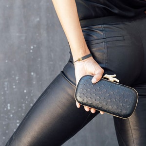 Puede incluir: Un clutch de cuero negro con un cierre dorado en forma de p&aacute;jaro. El clutch tiene un patr&oacute;n texturizado, similar al del avestruz.