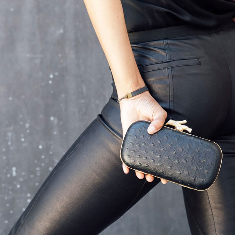 Puede incluir: Un clutch de cuero negro con un cierre dorado en forma de p&aacute;jaro. El clutch tiene un patr&oacute;n texturizado, similar al del avestruz.