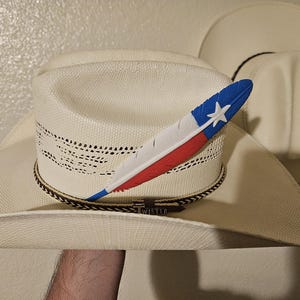 May include: A white straw cowboy hat featuring a woven texture, a brown and white patterned hatband with a metal Twister buckle, and a decorative feather accessory painted with blue, white, and red colors and a white star emblem on the side.