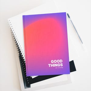 May include: A stack of notebooks with a pen. The top notebook has a gradient design of pink, red, and purple with the words "GOOD THINGS are coming" in white. The other notebooks are white and black.