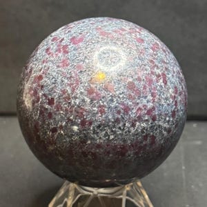 May include: A polished spherical stone featuring a mottled texture with deep red and dark grey crystalline inclusions throughout the surface. The sphere displays a reflective quality with light catching the varied mineral patterns across its round form.