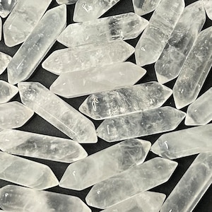 May include: A collection of clear quartz crystal points, also known as double terminated quartz, with a smooth, polished finish. The crystals are arranged in a random pattern on a black background.
