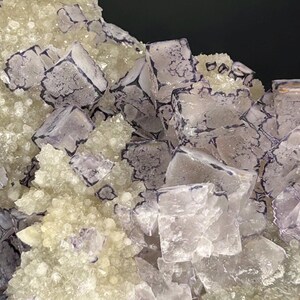 May include: Detailed image of a mineral specimen with cubic and crystalline structures. The translucent lavender crystals have dark purple edges, contrasted against a base of smaller, light-yellow crystals. The texture is rough and natural, showcasing geological formations.