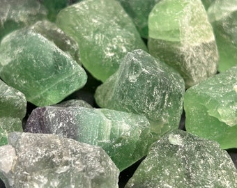 May include: A close-up of a pile of raw fluorite crystals. The crystals are a mix of green and purple hues, with a rough, textured appearance. The image highlights the natural, uncut state of the stones, showing their varied shapes and colours.