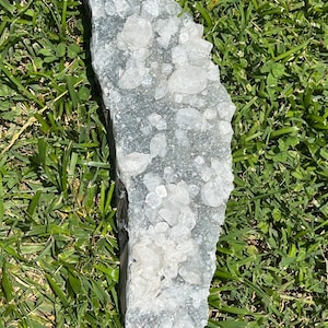 May include: A large, irregularly shaped crystal cluster with a light blue-gray hue. The surface is covered in numerous small, clear, crystalline formations. The crystal is resting on a bed of green grass.