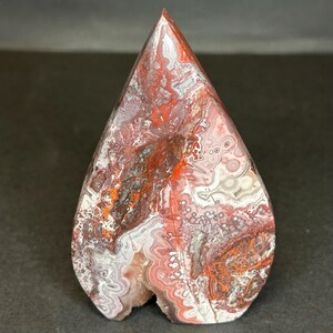 May include: A polished, teardrop-shaped gemstone with a pointed top. The stone features a complex pattern of red, white, and gray swirls and veins. The bottom of the stone has a small, natural cavity. The overall effect is a striking, natural art piece.