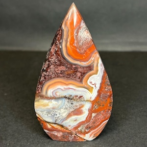 May include: A teardrop-shaped polished gemstone with a pointed top. The stone features layers of red, orange, white, and brown patterns. The gemstone has a smooth, glossy surface and is set against a dark background.