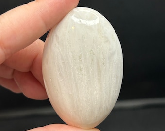 May include: A smooth, oval-shaped, white gemstone with subtle, light-colored veining. The stone is held between fingers, showcasing its polished surface and natural variations. The background is dark.