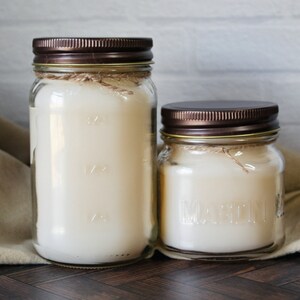 May include: Two glass candle jars with brown lids and a white candle inside. The larger jar has markings on the side.