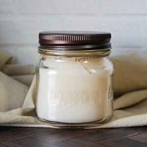 May include: A clear glass jar candle with a brown metal lid and a white wax candle inside. The jar has the word "MARTINI" embossed on the side.