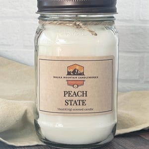 May include: A clear glass jar candle with a brown lid and a label that reads "Wauka Mountain Candleworks Since 2021 Peach State 16oz(454g) scented candle".