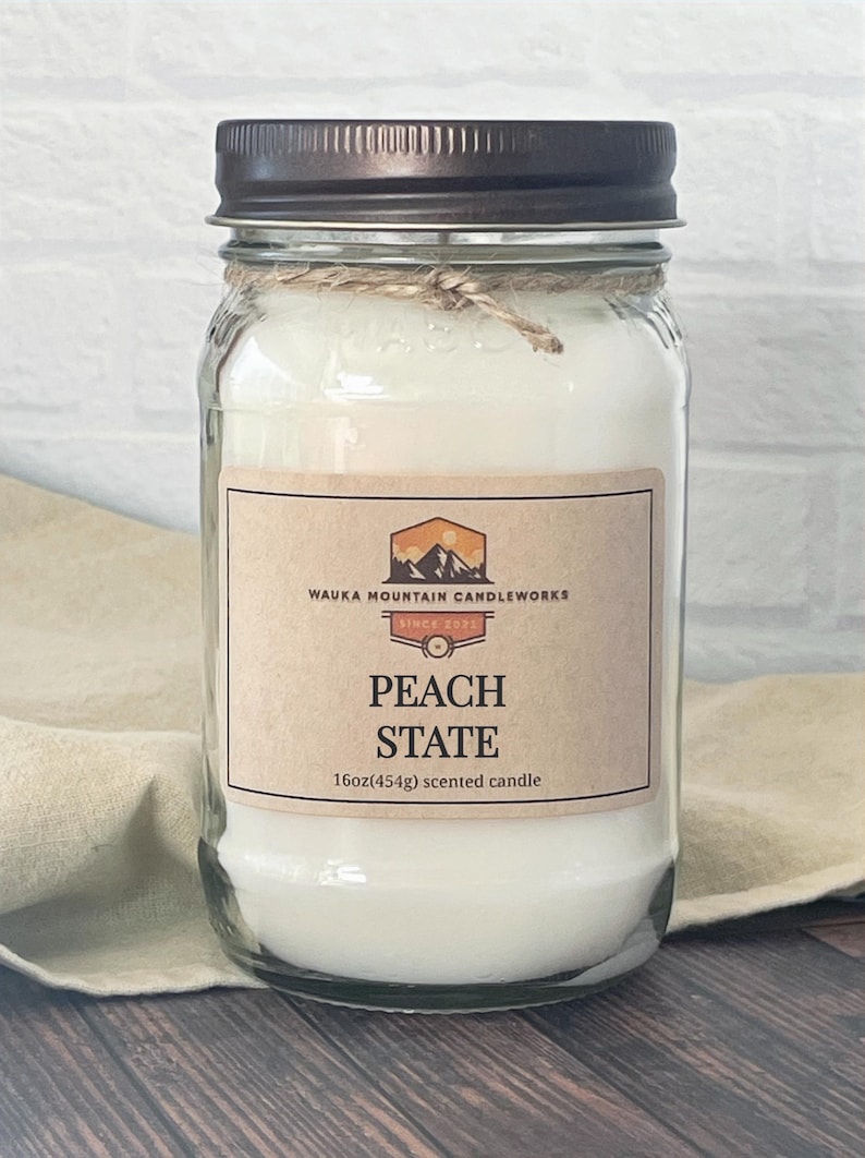May include: A clear glass jar candle with a brown lid and a label that reads "Wauka Mountain Candleworks Since 2021 Peach State 16oz(454g) scented candle".