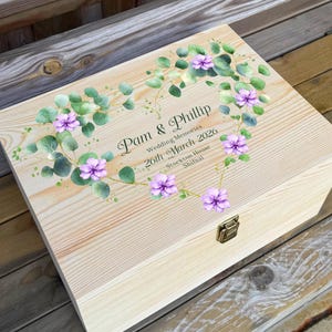 May include: A rectangular light-coloured wooden keepsake box with a metal latch. The lid features a printed heart-shaped wreath of green eucalyptus leaves and purple flowers, with the text Pam & Phillip, Wedding Memories, 26th March 2026, Stockton-House, Shifnal.