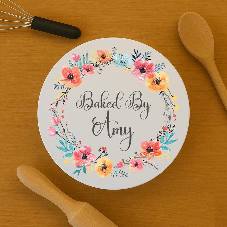 May include: A round white ceramic coaster featuring a floral wreath design in shades of pink, orange, and blue. The text "Baked By Amy" is printed in the center in a dark gray script font. A metal whisk and two wooden spoons are nearby.