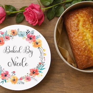 May include: A round white sticker featuring a floral wreath design in shades of pink, orange, and teal, with the text Baked By Nicole in a black script font, positioned next to a loaf of golden-brown cake inside a round container.