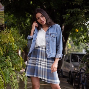 May include: A woman wearing a blue and white plaid skirt, a white shirt, and a blue denim jacket. She is standing in front of a green leafy plant.