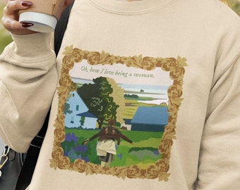 May include: A beige sweatshirt with a graphic print of a house and a person in a field. The text "Oh, how I love being a woman." is printed on the graphic.