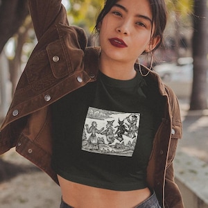 May include: A black crop top with a vintage illustration of a group of people dancing with a devil. The top is worn by a person with brown hair and a brown leather jacket.