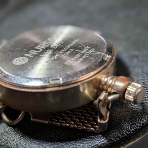 May include: A silver-toned watch case back with a metallic finish. The text reads NURSE, NMS-2475, WATER RESISTANT 10BAR, SS case back, and Made in China. A textured metal mesh strap is attached to the side, along with a small winding crown.